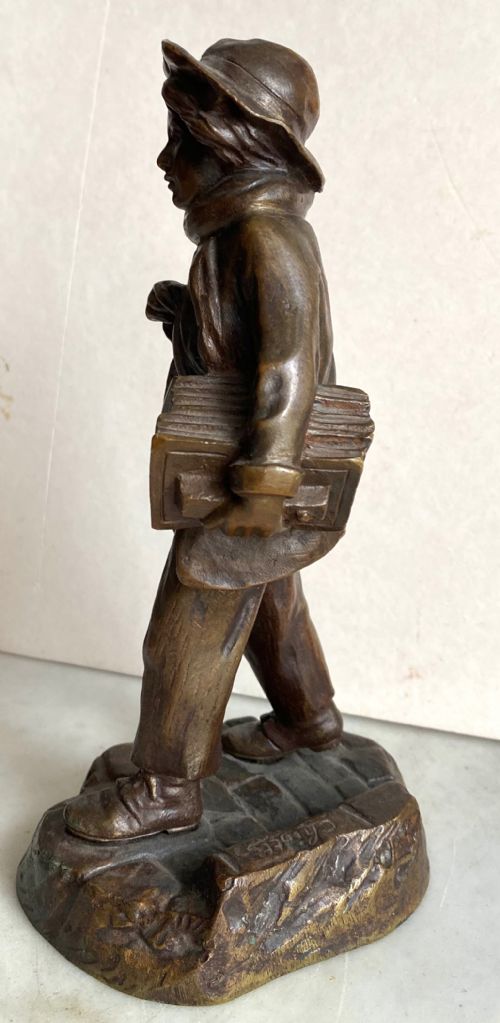fig 3. A 19th-century patinated bronze sculpture depicting a schoolboy in a coat, cap, and scarf carrying a strap of books across his shoulder, standing on a cobblestone base.
