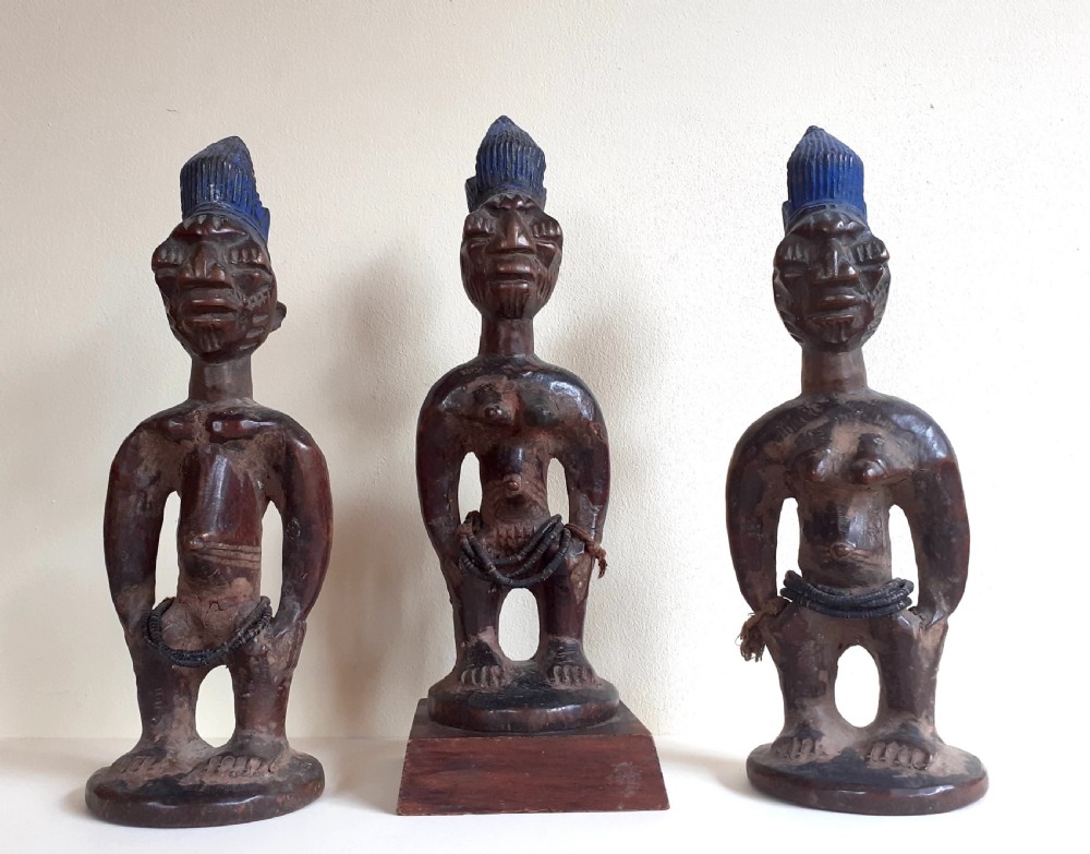 Three hand-carved dark wood Yoruba Ere Ibeji figures featuring tall indigo-pigmented coiffures, carved facial scarification, and glass waist beads.