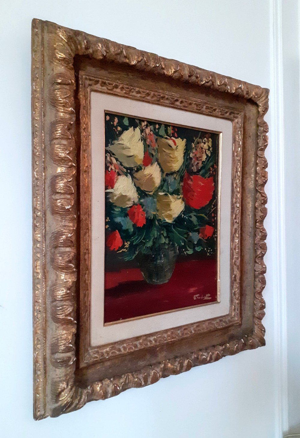 A framed oil painting on canvas depicting a bouquet of yellow and red flowers in a glass vase set on a dark red table surface. View V
