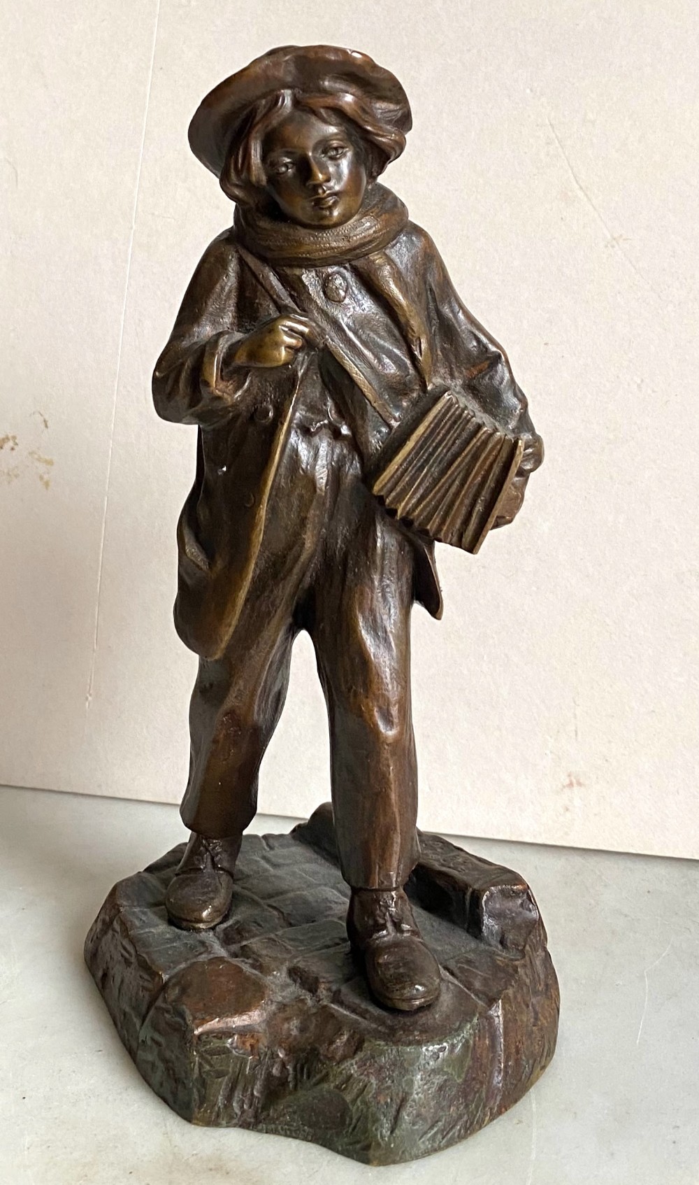 A 19th-century patinated bronze sculpture depicting a schoolboy in a coat, cap, and scarf carrying a strap of books across his shoulder, standing on a cobblestone base.
