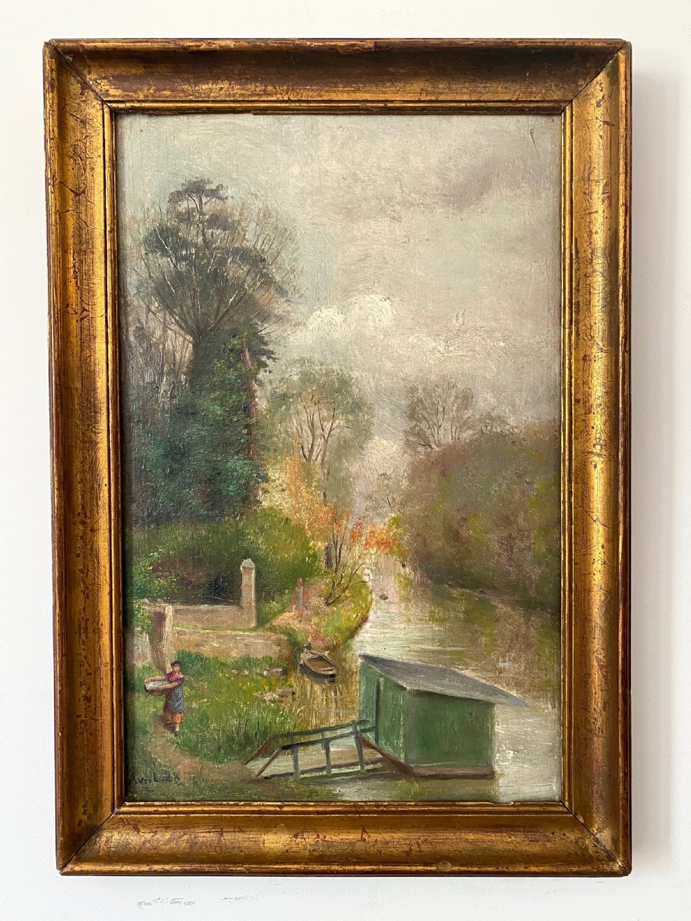 An antique French oil painting on a wooden panel depicting a riverbank scene with a figure carrying a basket, trees, and a green structure under an overcast sky. View II