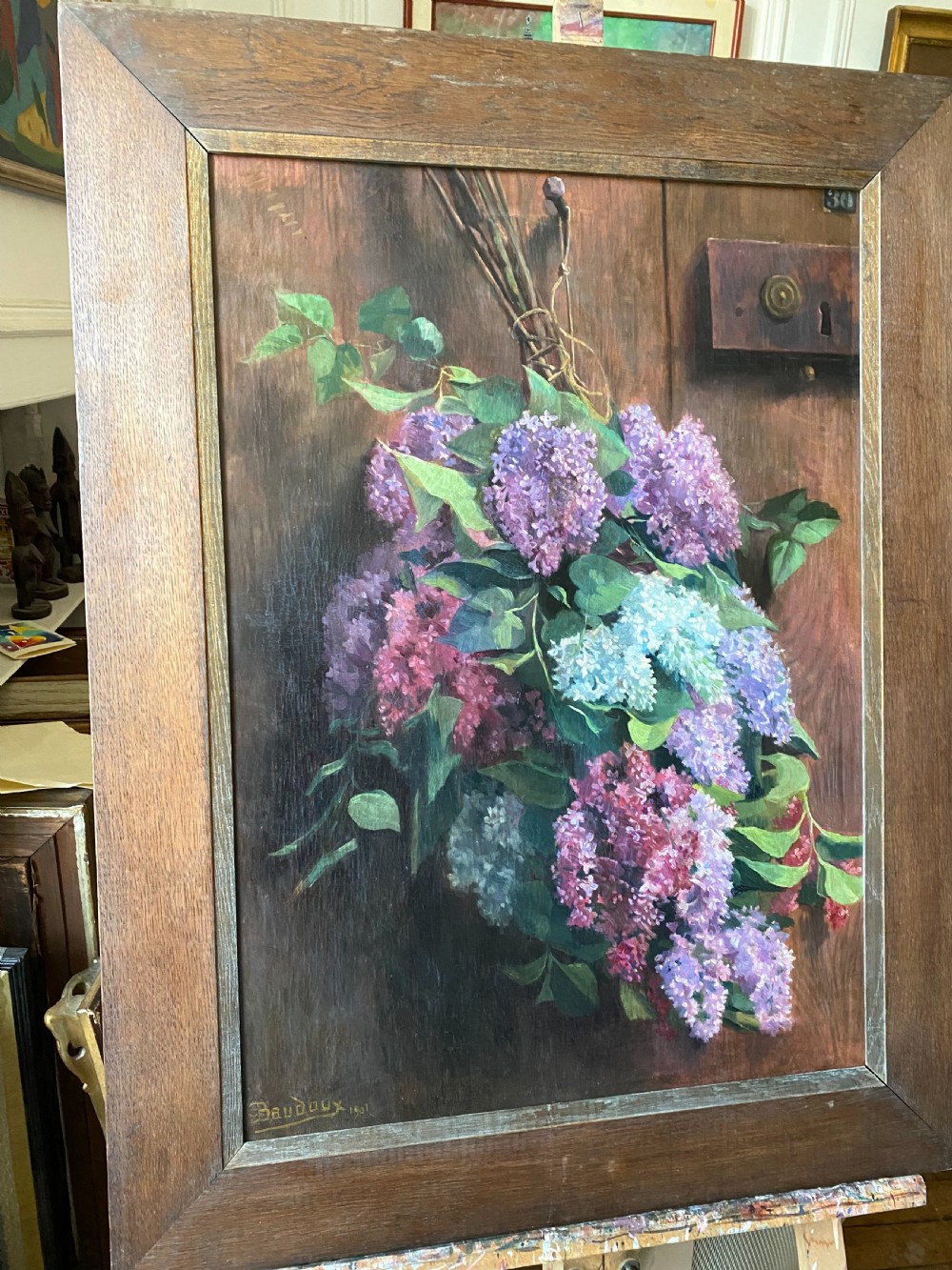 An oil on canvas painting dated 1901 depicting a bouquet of purple and white lilac flowers hanging against a dark oak wooden door. View VI