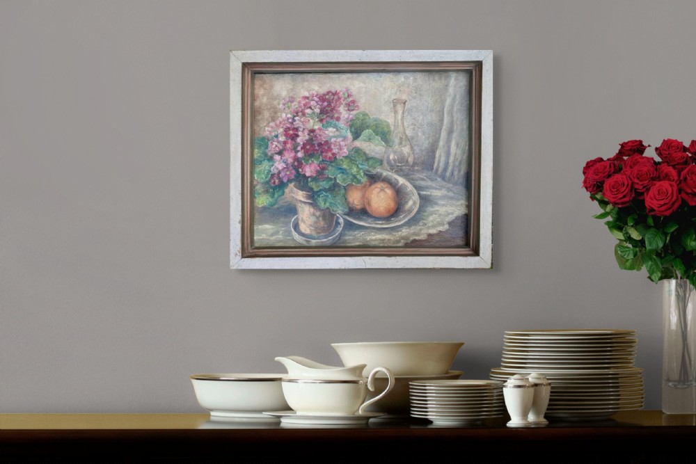 A framed 1934 still life oil painting on canvas depicting a potted pink geranium, two oranges in a dish, and a glass vessel on a table. View III