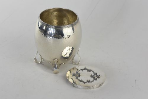 fig 2. An Arts and Crafts hammered sterling silver tea caddy featuring a planished surface, looped feet, and a flat lid with a scrolled handle.