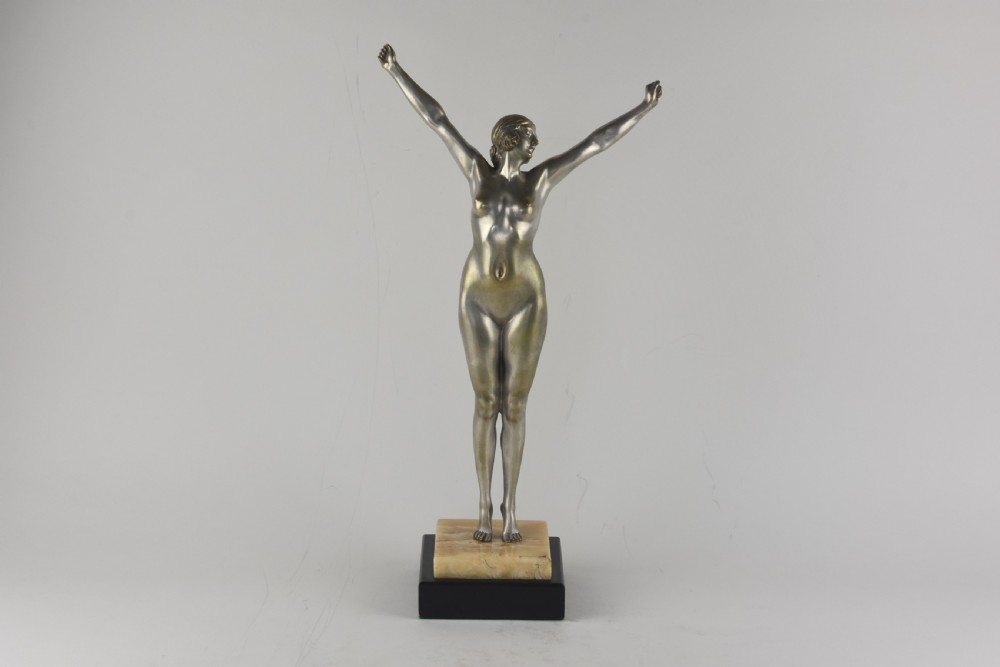 An Art Deco silvered bronze sculpture depicting a nude female figure with raised arms standing atop a tiered marble and black base.