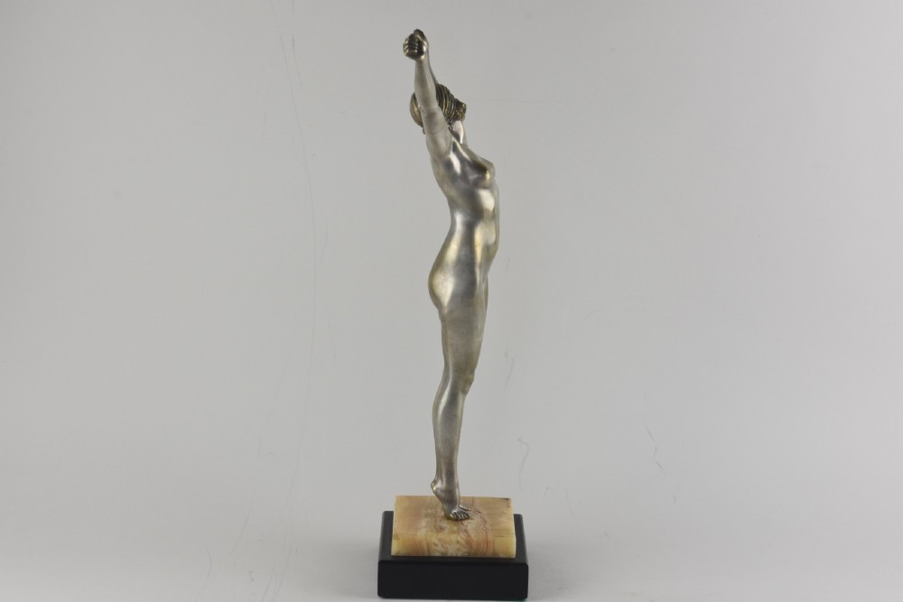An Art Deco silvered bronze sculpture depicting a nude female figure with raised arms standing atop a tiered marble and black base. View VI