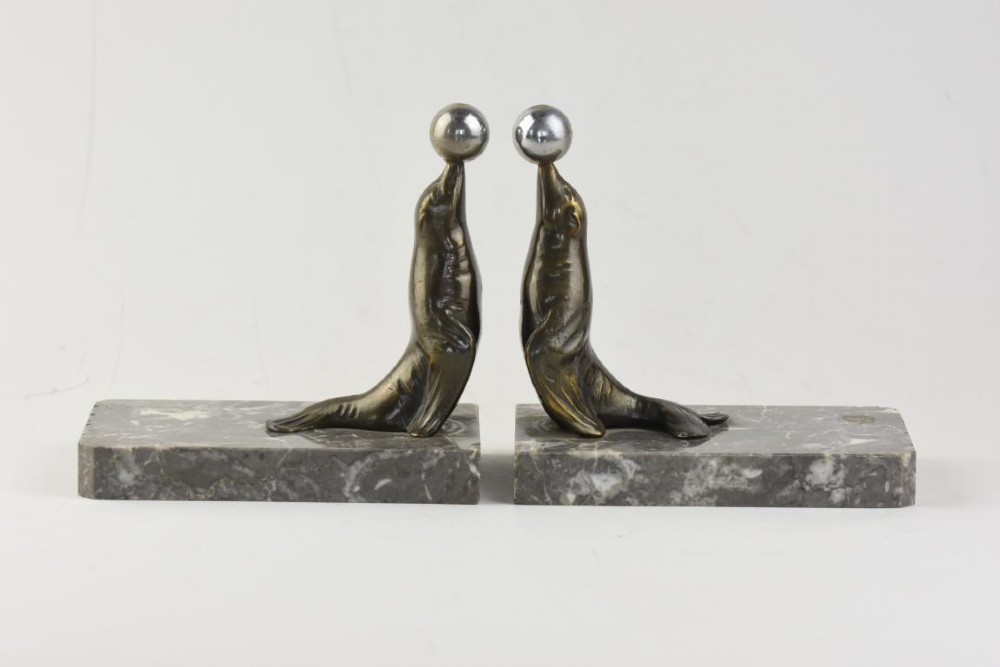A pair of Art Deco metal sea lions balancing chrome spheres on their noses, mounted on rectangular grey marble bases.