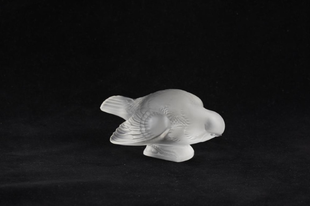 A clear frosted glass sculpture depicting a small sparrow with its head lowered in a shy pose against a black background.