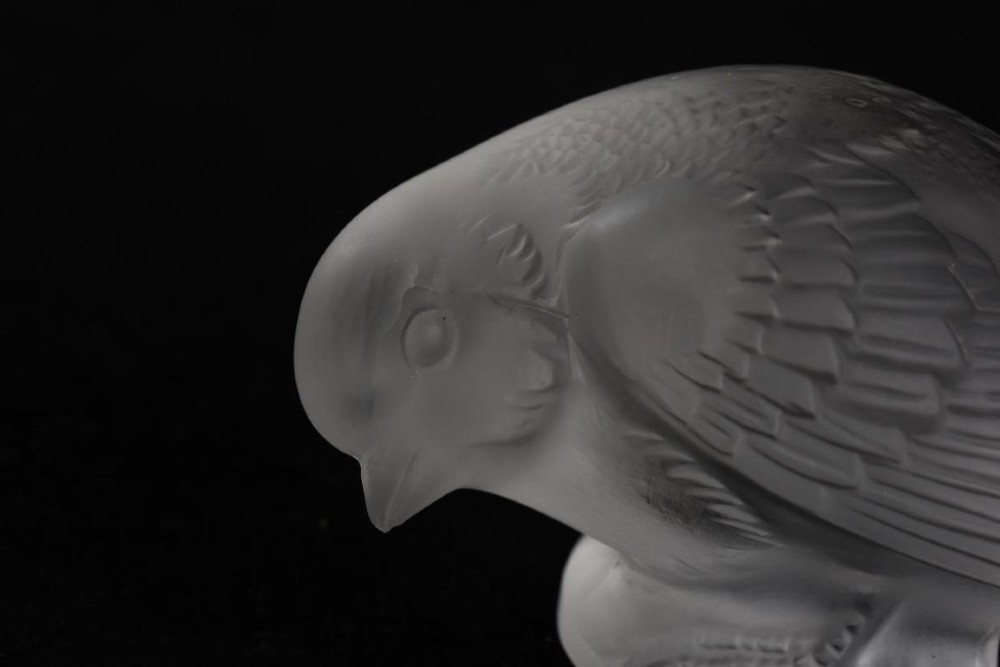A clear frosted glass sculpture depicting a small sparrow with its head lowered in a shy pose against a black background. View III