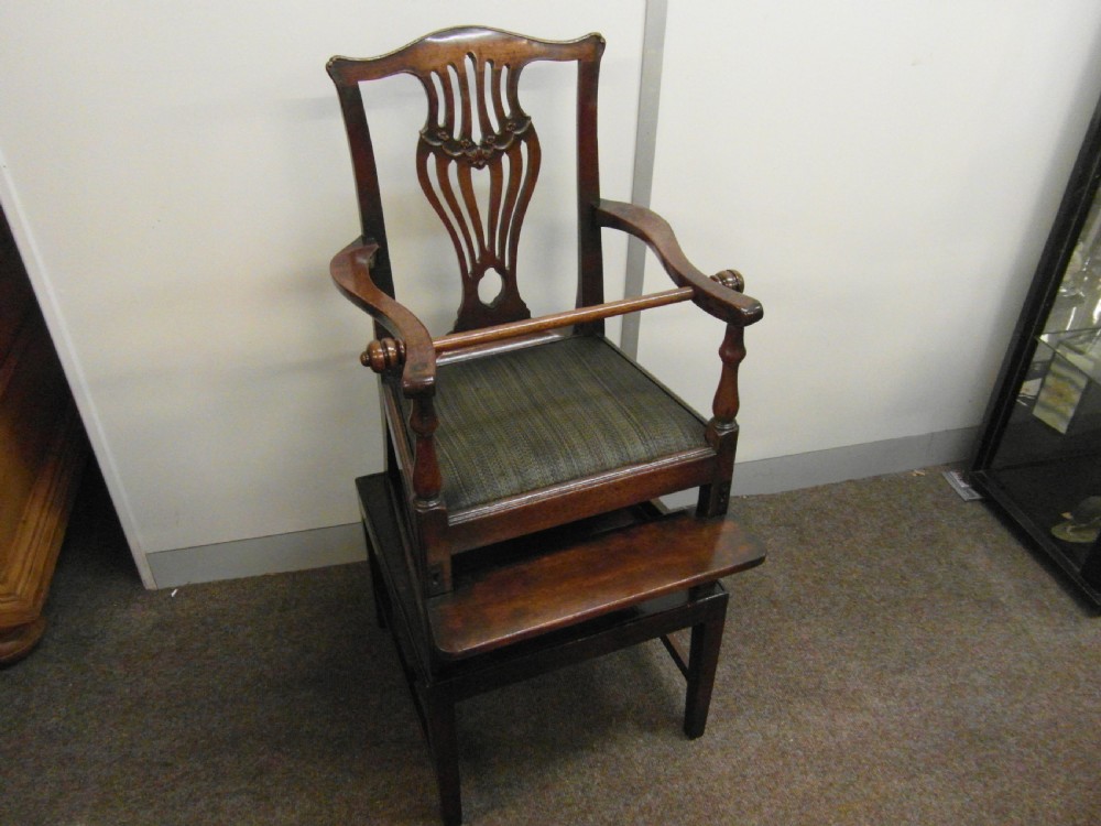 An antique Georgian mahogany childs high chair featuring a carved pierced splat back, wooden safety bar, upholstered seat, and integrated footrest on a wooden stand.