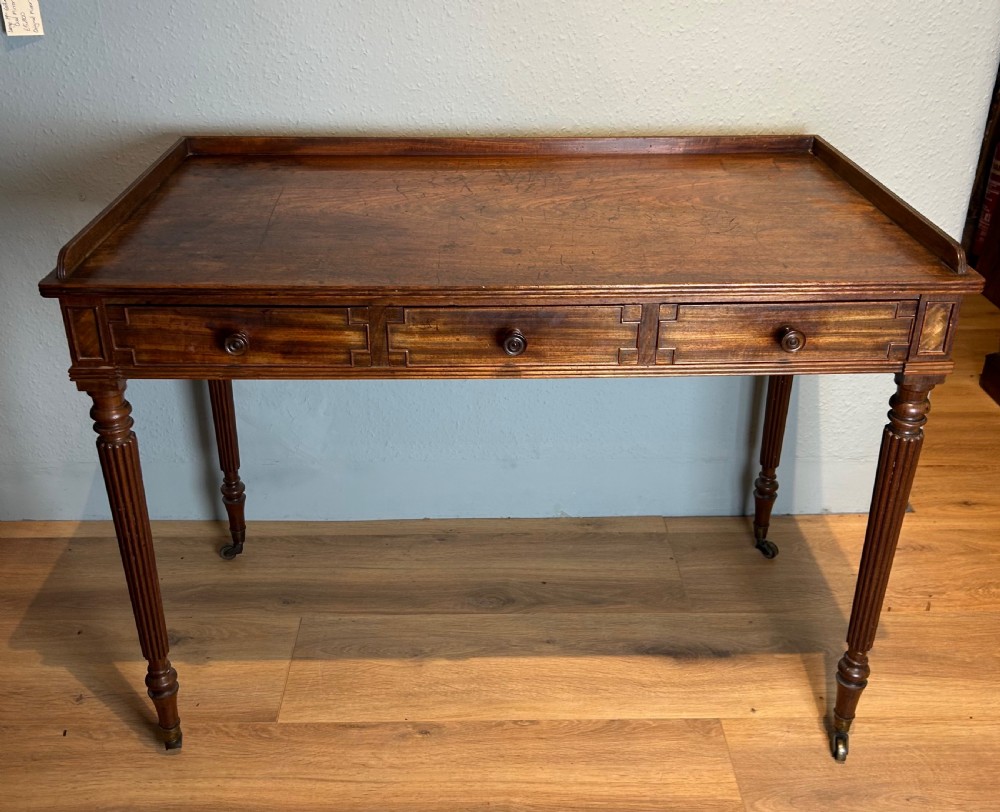 A Regency mahogany writing table featuring a gallery top, three frieze drawers with turned knob handles, and slender reeded turned legs on brass castors.