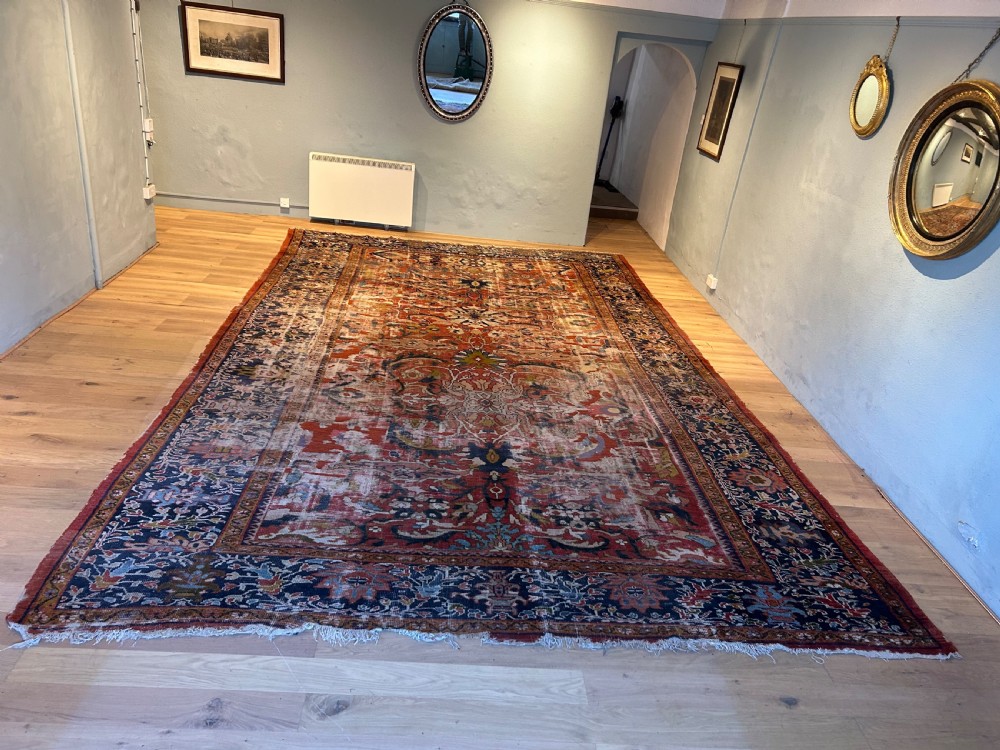 A large hand-knotted antique Ziegler carpet featuring floral palmette patterns in terracotta red, navy blue, and cream tones laid flat on a wooden floor.
