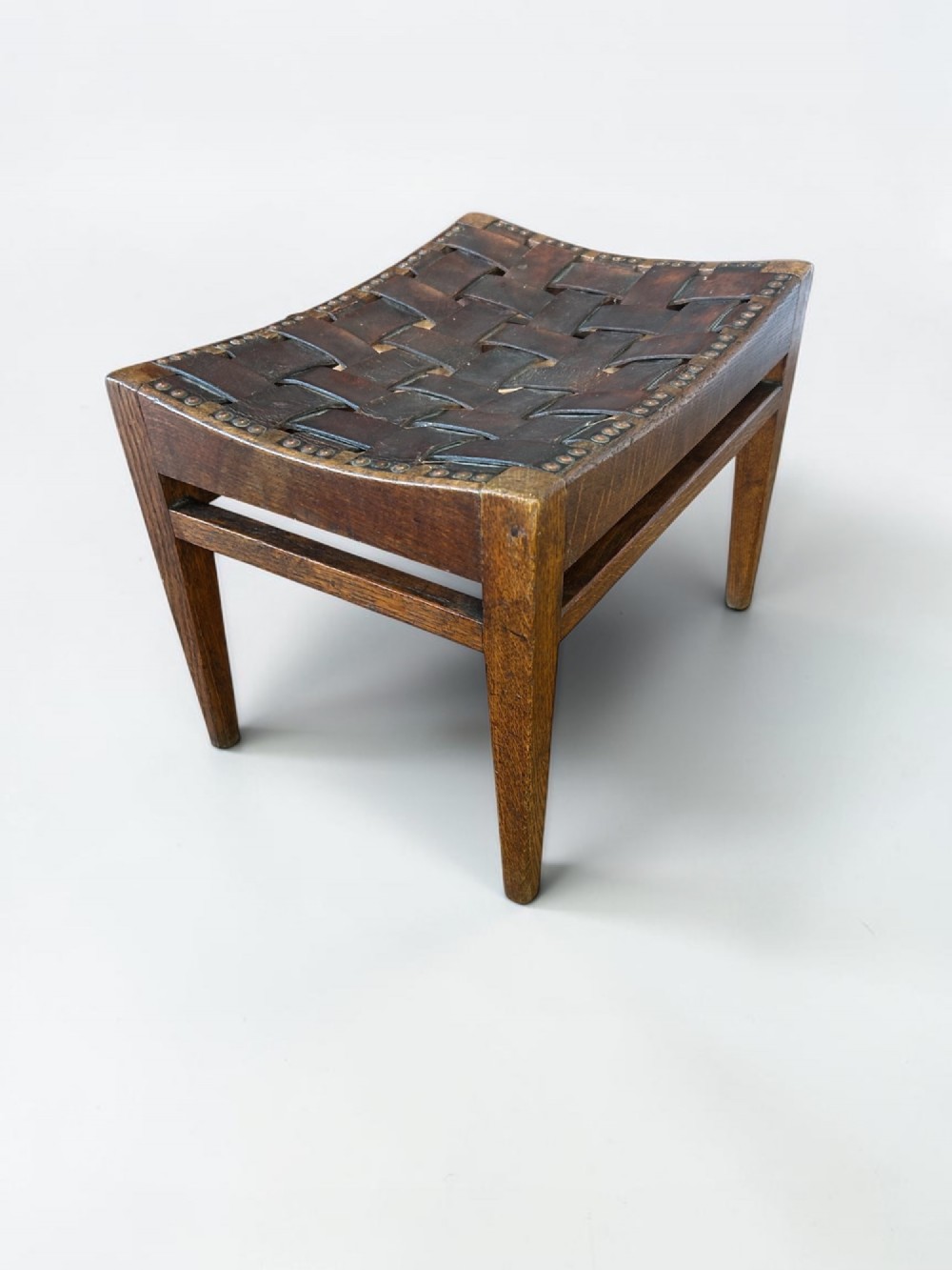 An Arts and Crafts dark oak stool featuring a concave woven leather strap seat secured with brass studs on tapered square legs.