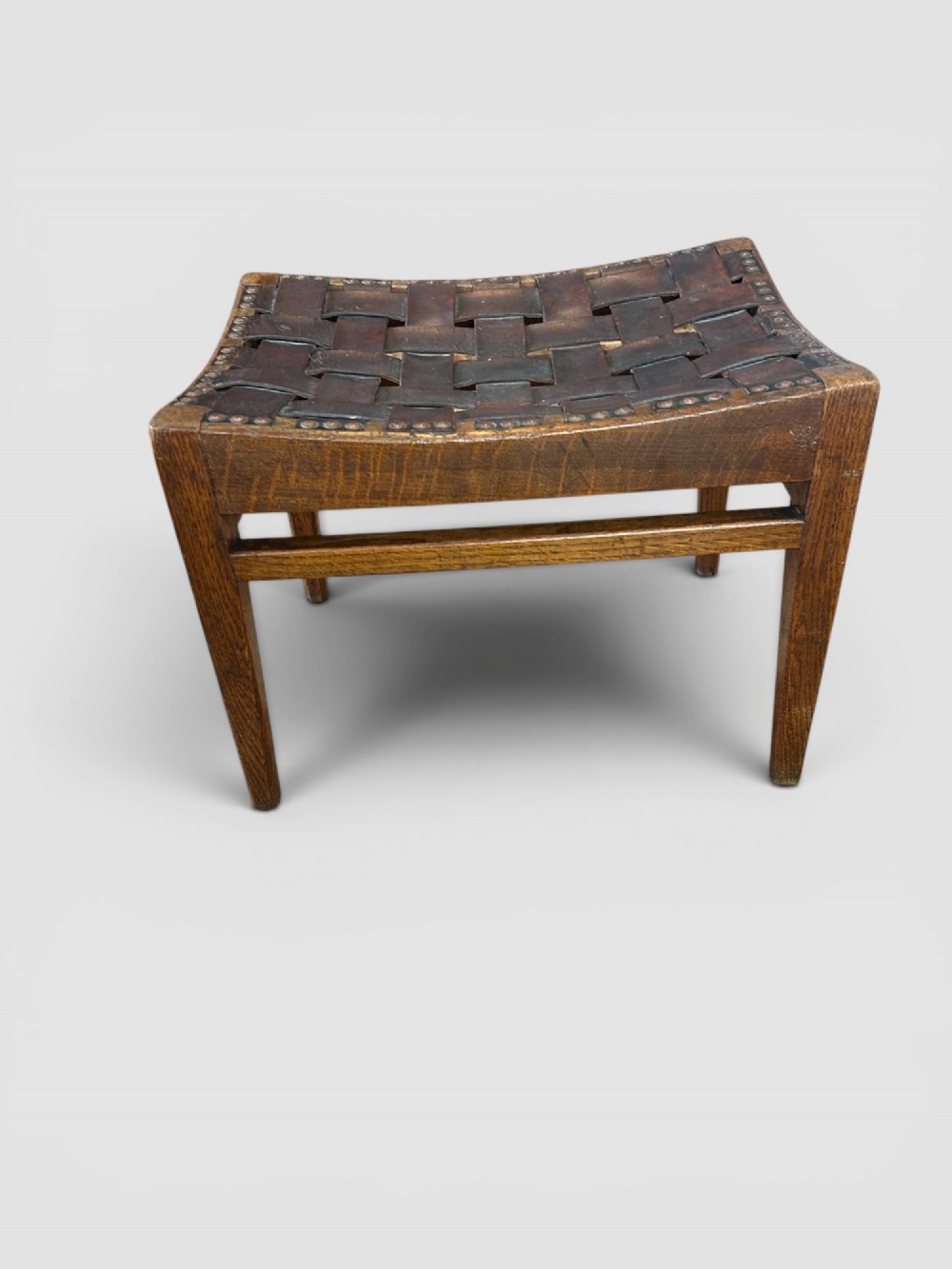 An Arts and Crafts dark oak stool featuring a concave woven leather strap seat secured with brass studs on tapered square legs. View II