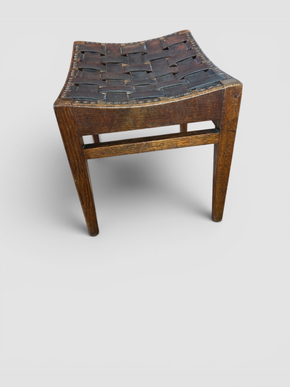 An Arts and Crafts dark oak stool featuring a concave woven leather strap seat secured with brass studs on tapered square legs. View III