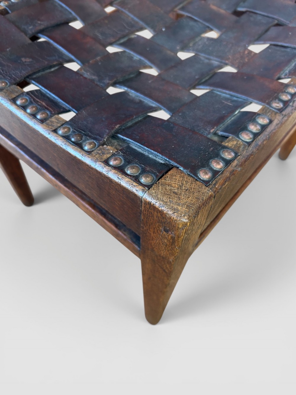 An Arts and Crafts dark oak stool featuring a concave woven leather strap seat secured with brass studs on tapered square legs. View V
