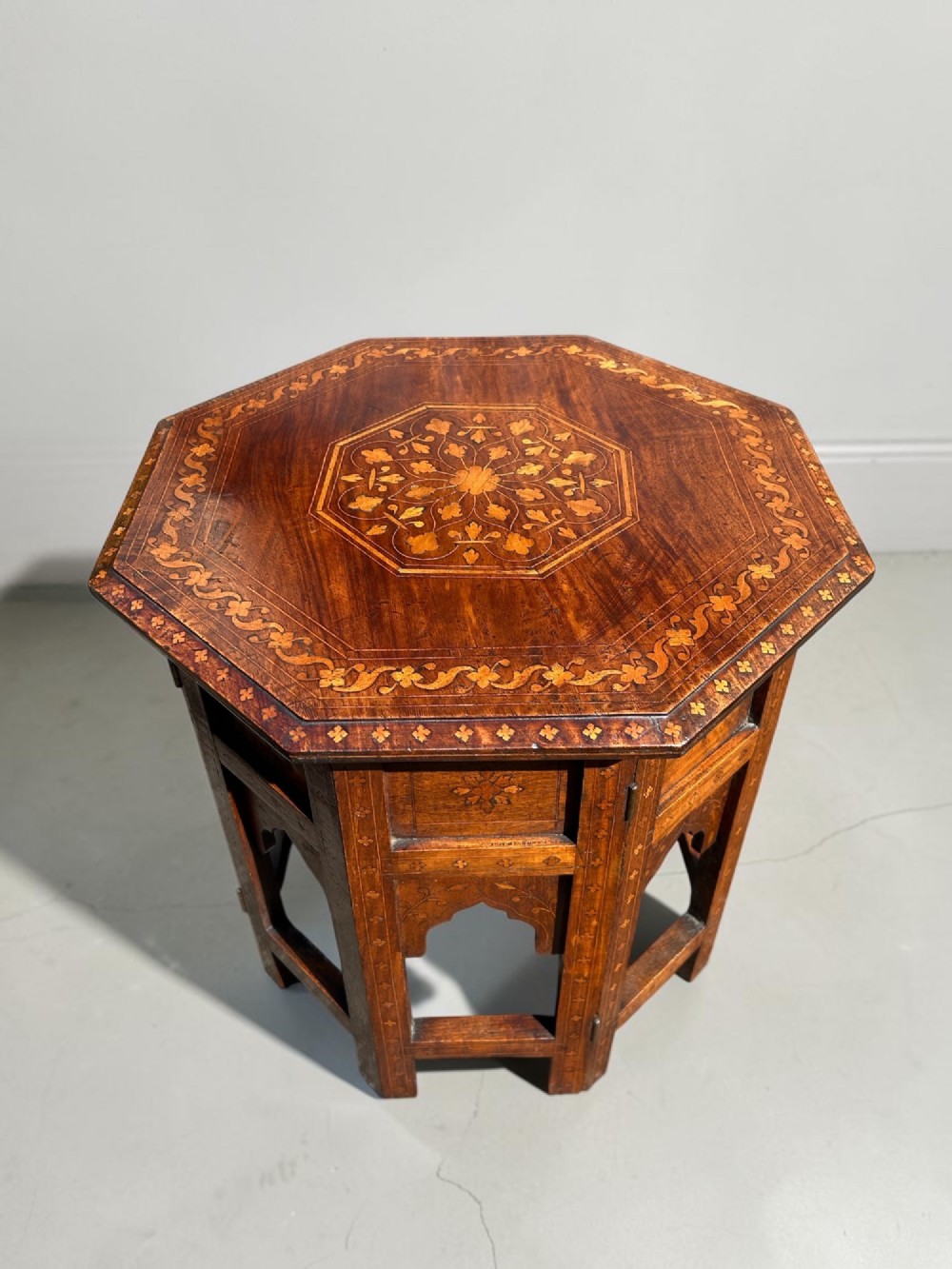 An octagonal Anglo-Indian wooden side table featuring intricate floral marquetry inlay across the top and an openwork arched apron base.