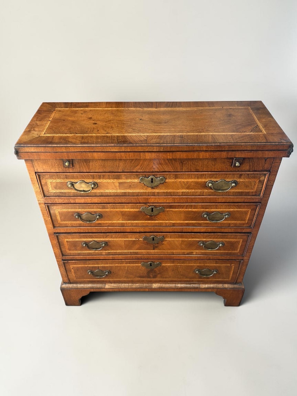 A George I walnut bachelors chest featuring four graduated drawers with feather-banded borders, brass bail handles, keyhole escutcheons, and shaped bracket feet. View II