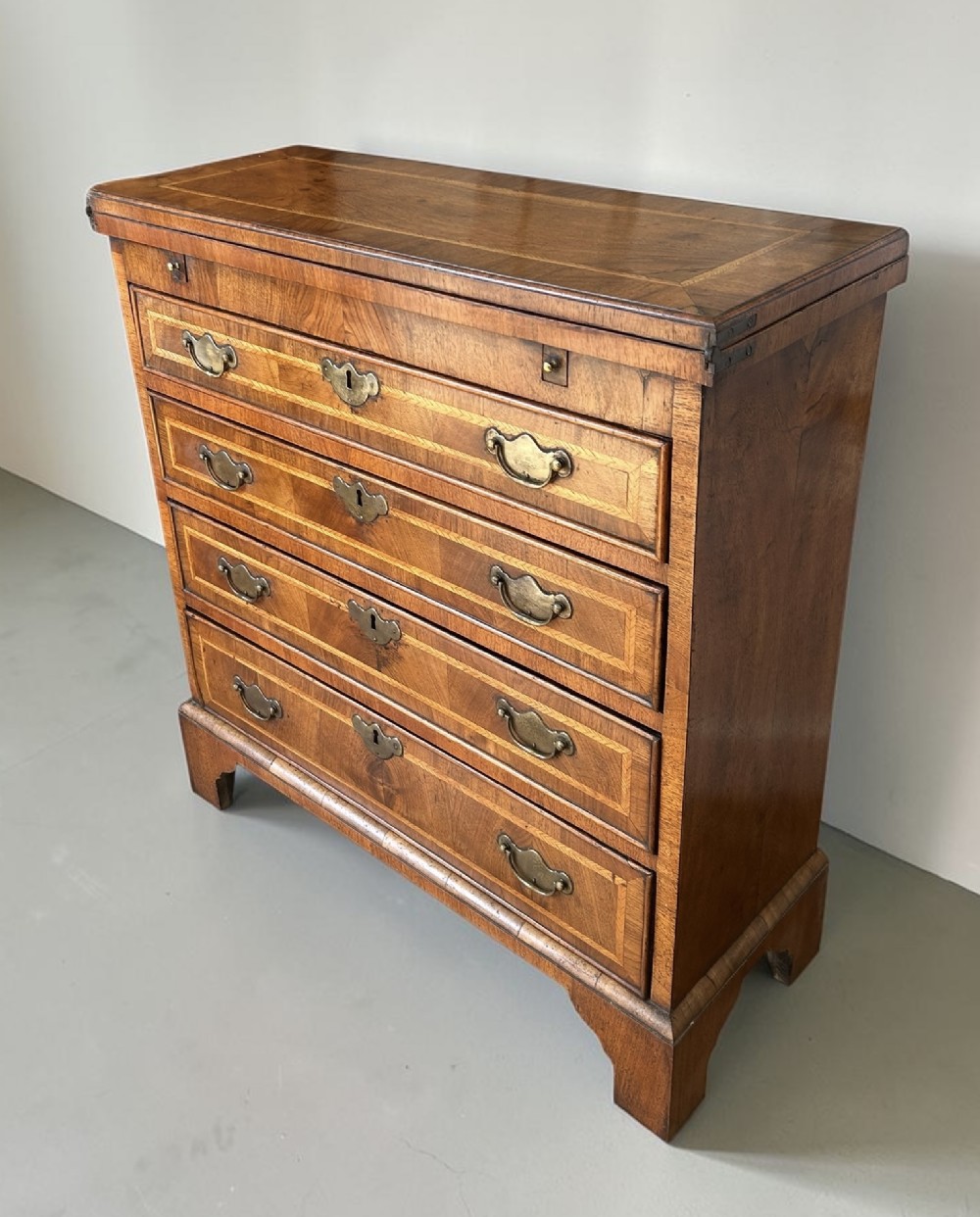 A George I walnut bachelors chest featuring four graduated drawers with feather-banded borders, brass bail handles, keyhole escutcheons, and shaped bracket feet.
