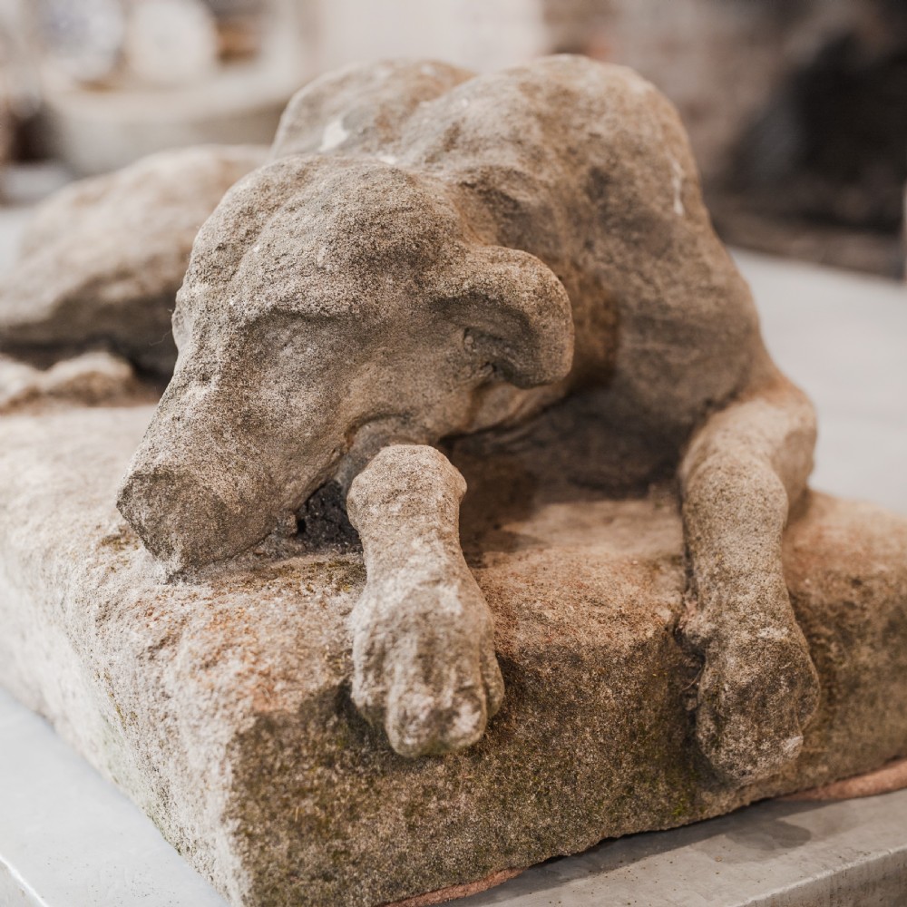 A weathered reconstituted stone sculpture depicting a recumbent hound resting its head on its paws atop a rectangular base.