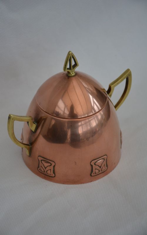 fig 1. An Art Nouveau copper sugar bowl featuring angular brass handles, a conical lid with a stylised finial, and embossed geometric motifs around the base.
