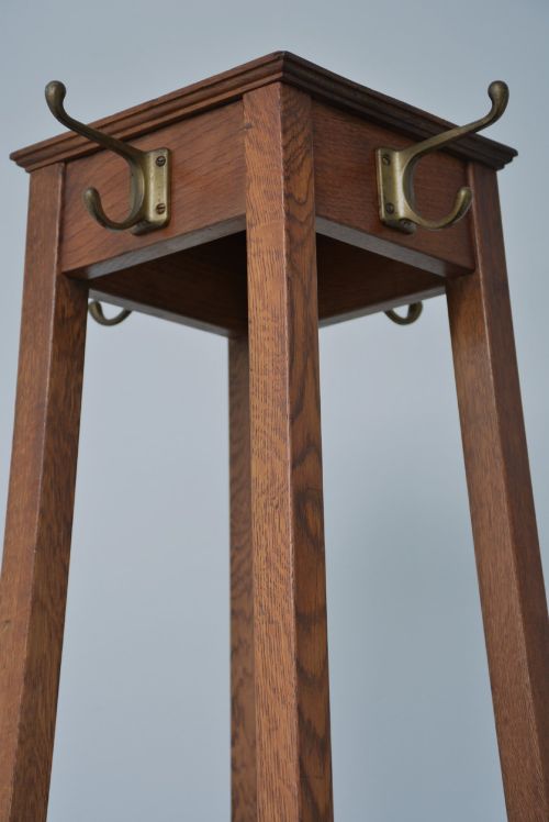 fig 1. A tall, tapered solid oak Arts and Crafts coat stand featuring brass hooks at the top, a middle support bar, and a square drip tray at the base.