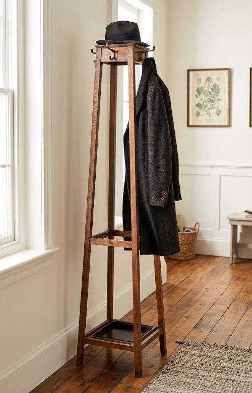 A tall, tapered solid oak Arts and Crafts coat stand featuring brass hooks at the top, a middle support bar, and a square drip tray at the base. View XIV
