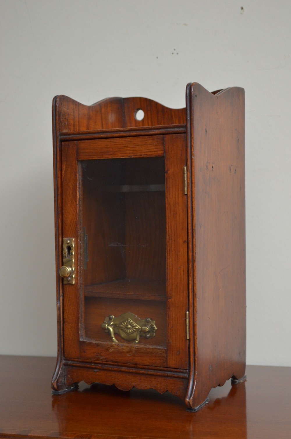 An Arts and Crafts oak smokers cabinet featuring a single glazed door, decorative brass handles, and a shaped top gallery with a central circular cutout.