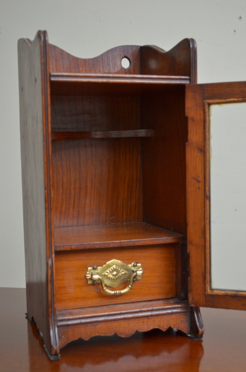 An Arts and Crafts oak smokers cabinet featuring a single glazed door, decorative brass handles, and a shaped top gallery with a central circular cutout. View II