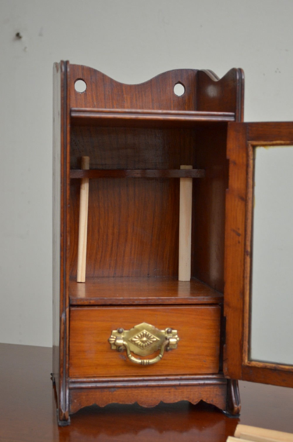 An Arts and Crafts oak smokers cabinet featuring a single glazed door, decorative brass handles, and a shaped top gallery with a central circular cutout. View III