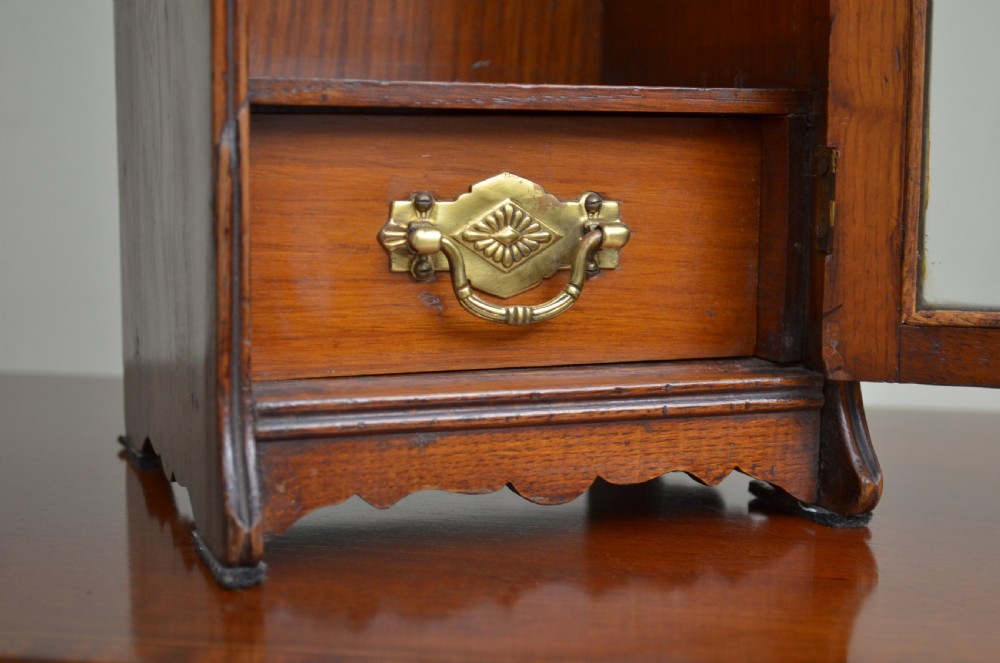 An Arts and Crafts oak smokers cabinet featuring a single glazed door, decorative brass handles, and a shaped top gallery with a central circular cutout. View IV