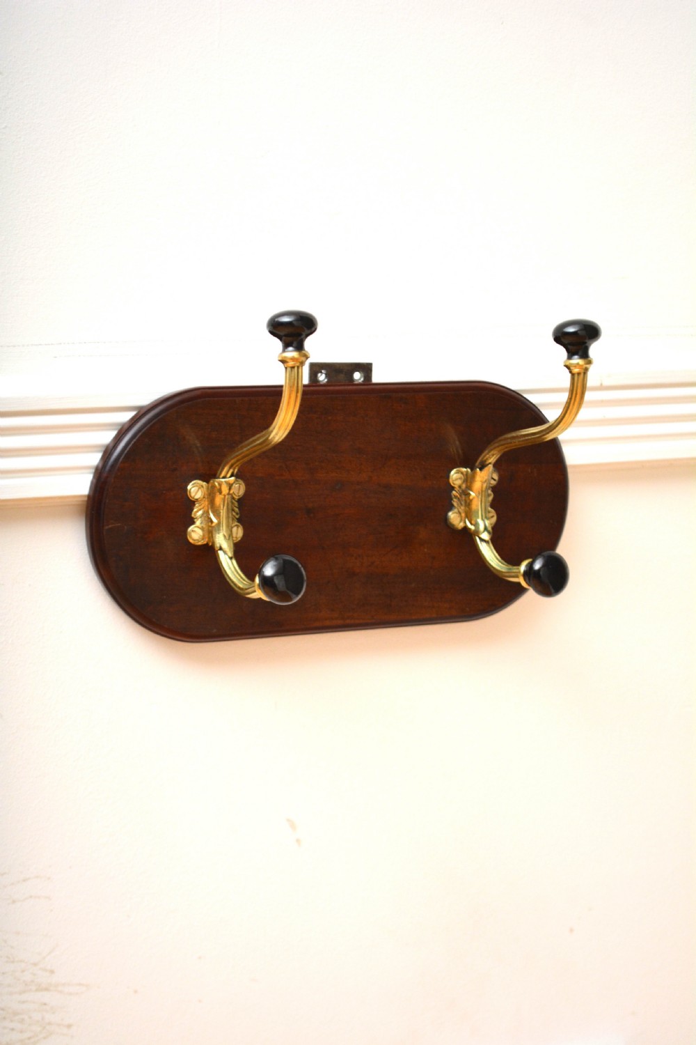 An antique wall-mounted coat rack featuring a polished mahogany backboard fitted with two ornate brass hooks with black ceramic finials.