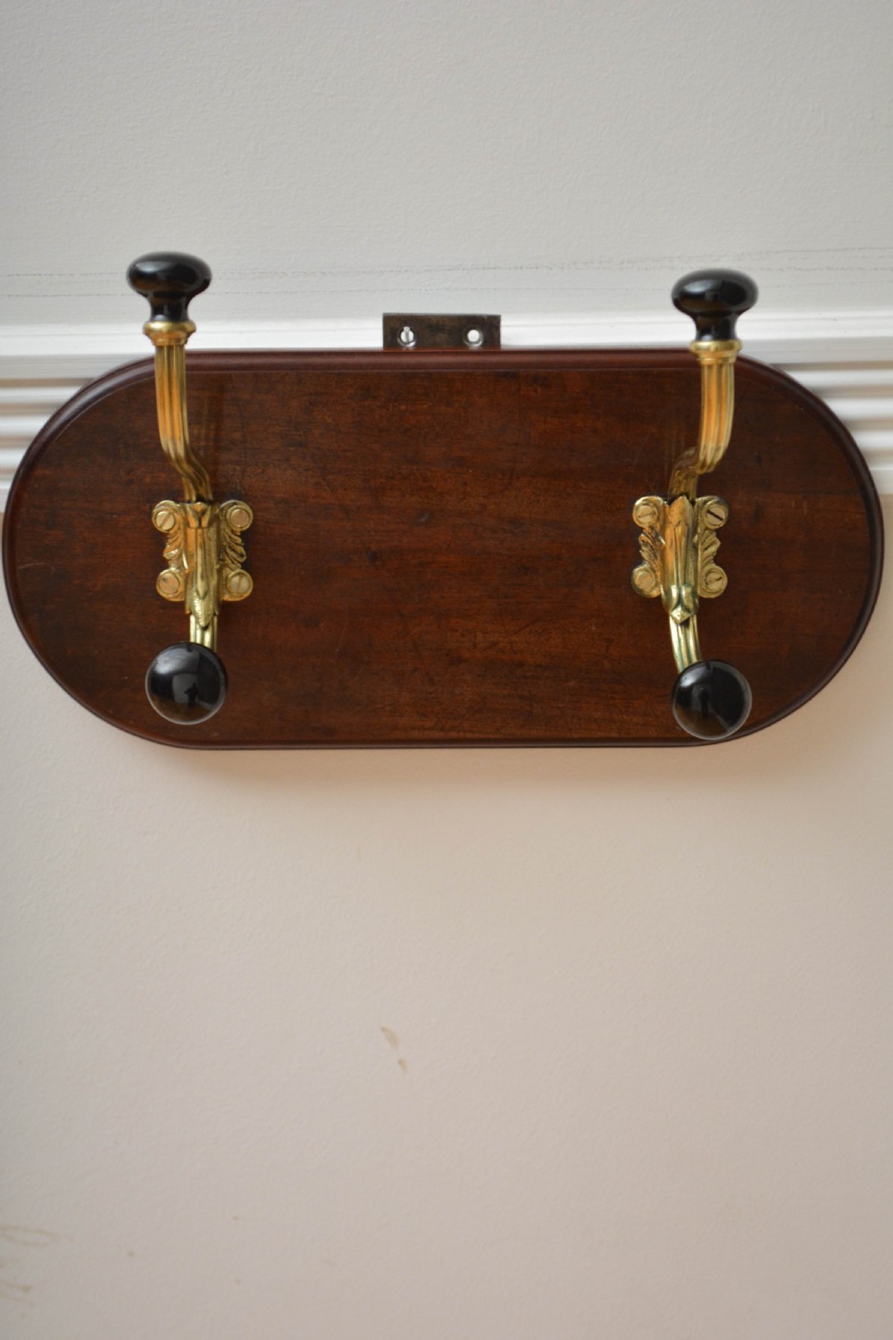 An antique wall-mounted coat rack featuring a polished mahogany backboard fitted with two ornate brass hooks with black ceramic finials. View II