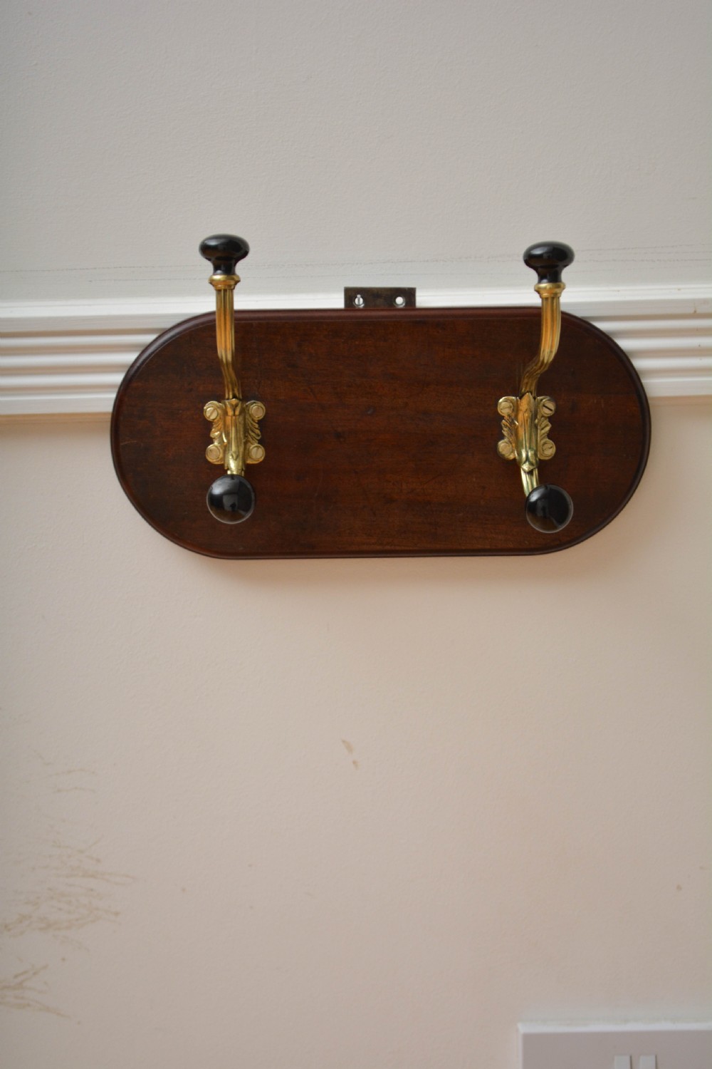 An antique wall-mounted coat rack featuring a polished mahogany backboard fitted with two ornate brass hooks with black ceramic finials. View III
