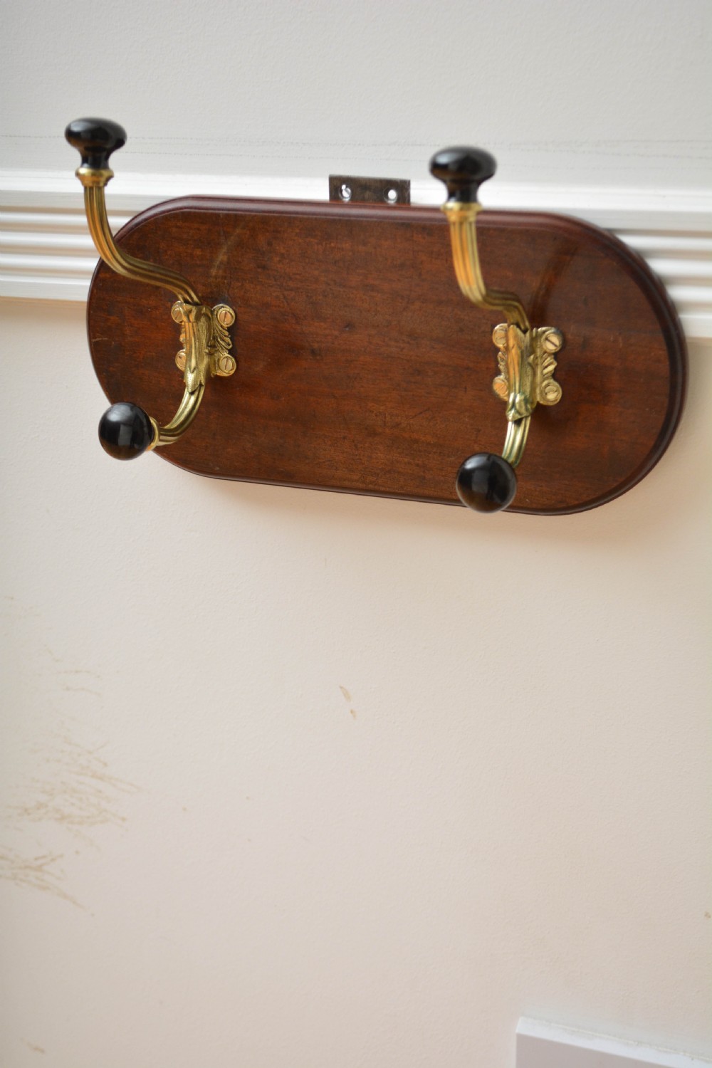 An antique wall-mounted coat rack featuring a polished mahogany backboard fitted with two ornate brass hooks with black ceramic finials. View V