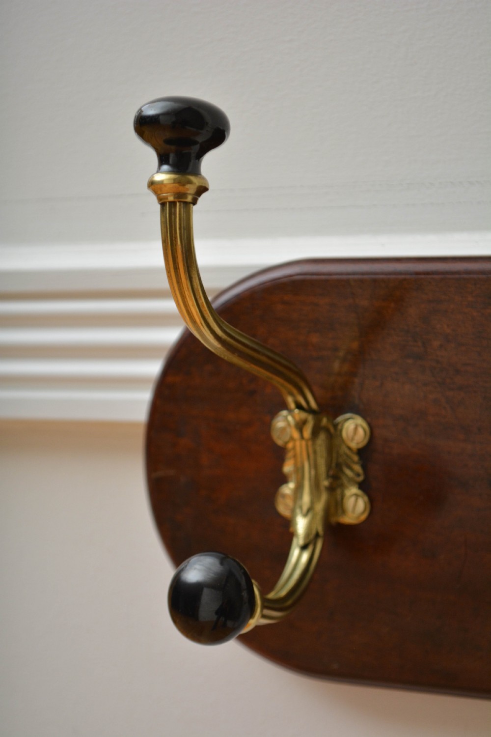 An antique wall-mounted coat rack featuring a polished mahogany backboard fitted with two ornate brass hooks with black ceramic finials. View VI