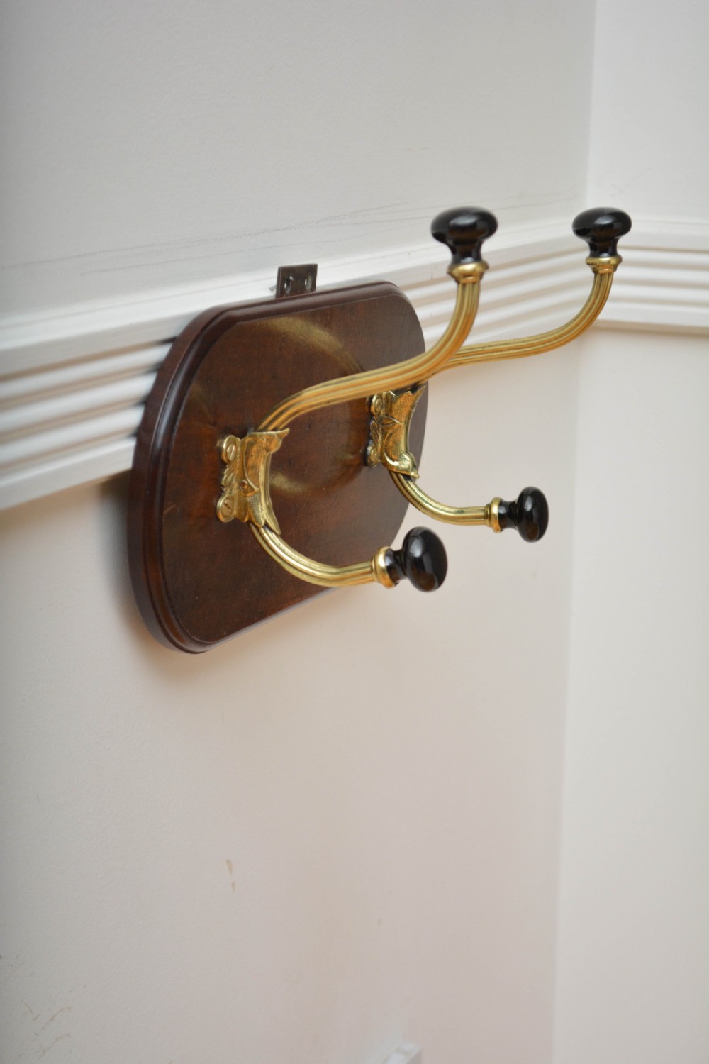 An antique wall-mounted coat rack featuring a polished mahogany backboard fitted with two ornate brass hooks with black ceramic finials. View VII