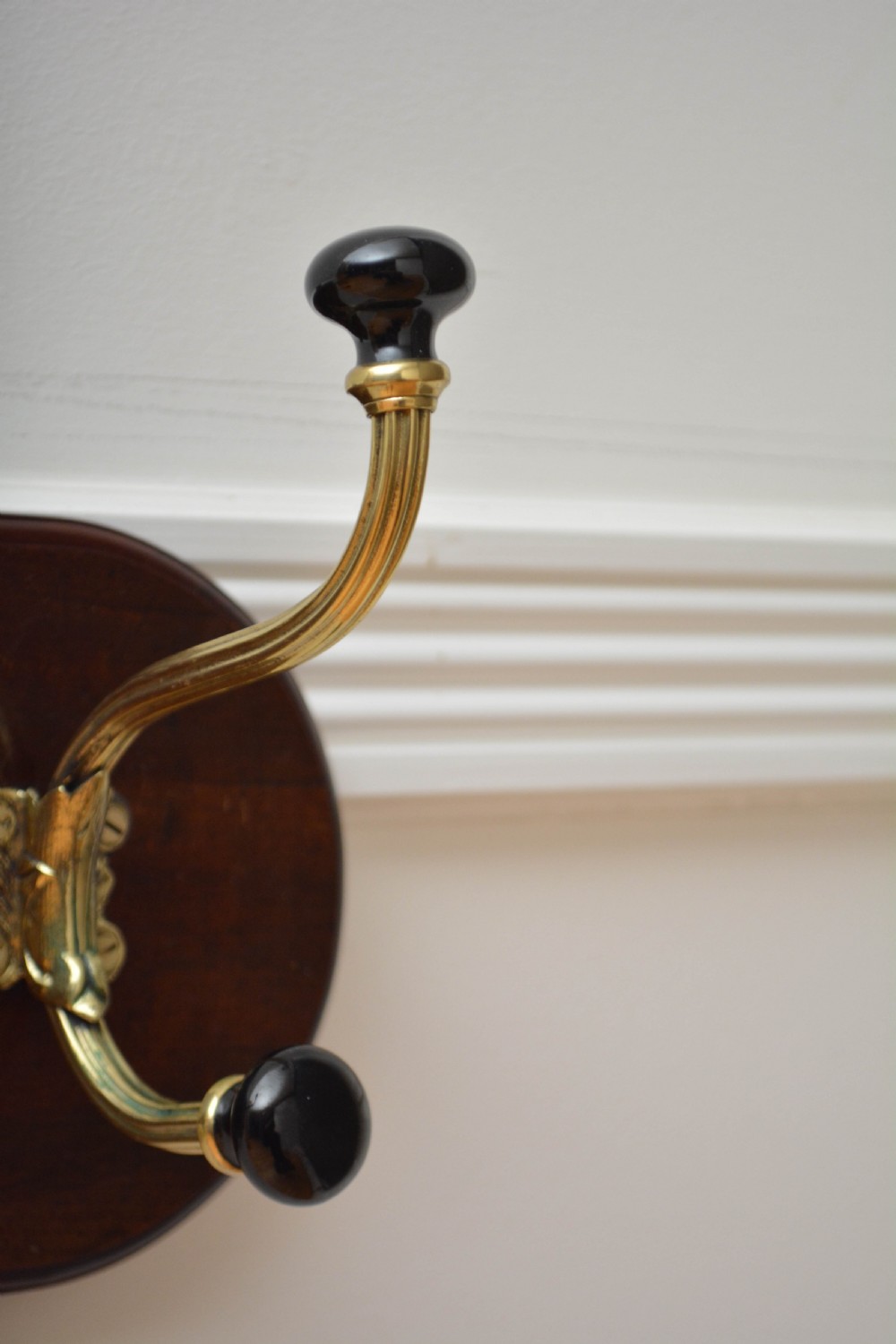 An antique wall-mounted coat rack featuring a polished mahogany backboard fitted with two ornate brass hooks with black ceramic finials. View IX