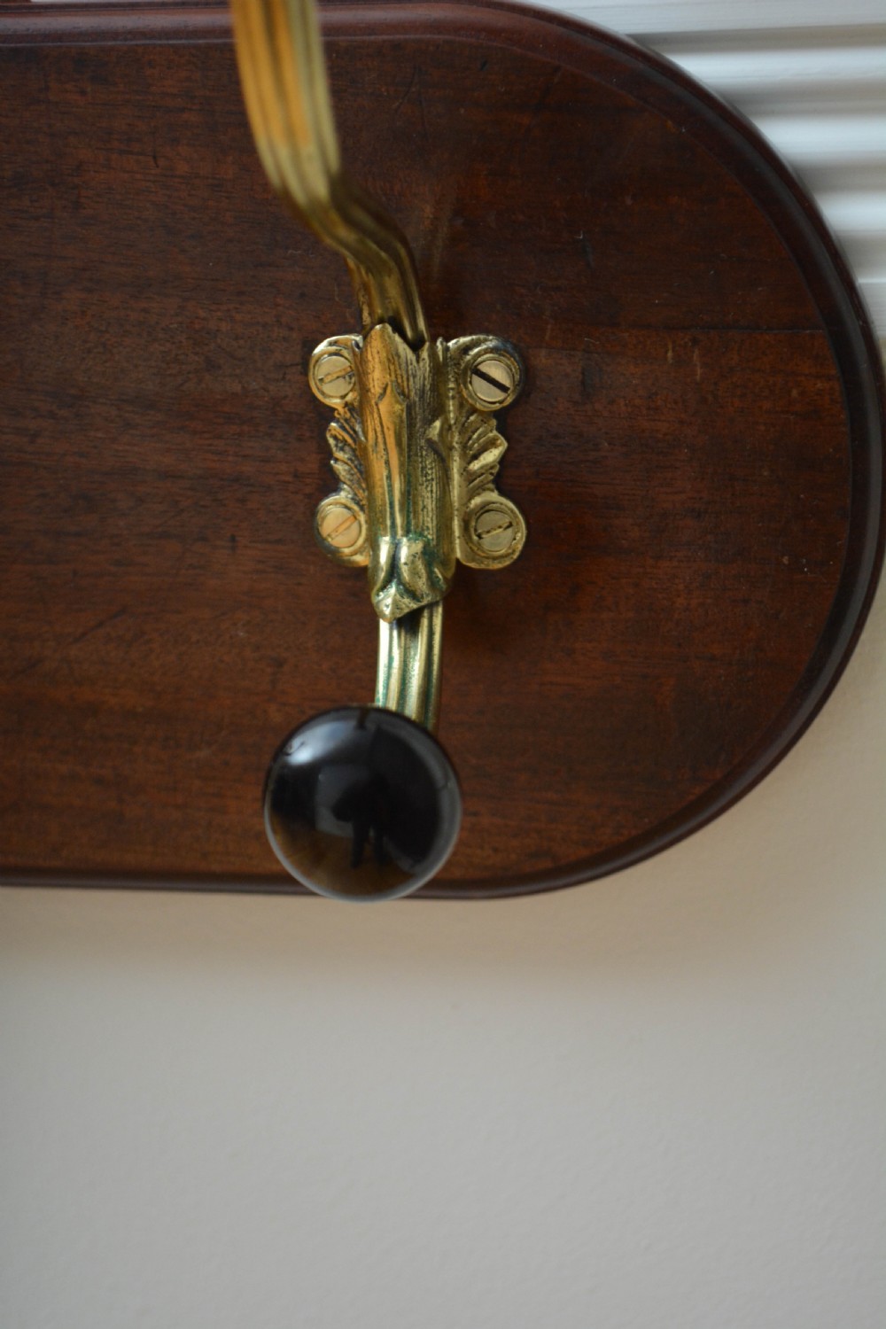 An antique wall-mounted coat rack featuring a polished mahogany backboard fitted with two ornate brass hooks with black ceramic finials. View XII