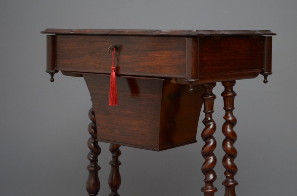 A Victorian rosewood work table featuring a hinged top surface, a keyhole drawer, a lower wooden work basket, four barley twist support legs, and scroll feet. View IX