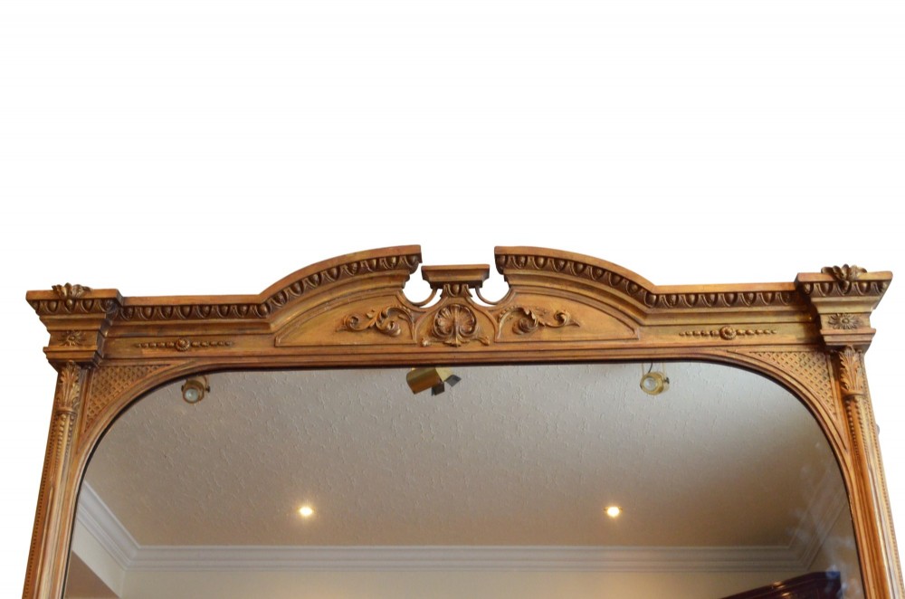 A large rectangular giltwood overmantel mirror featuring an arched top frame, carved cresting, side pilasters, and a gold finish. View VI