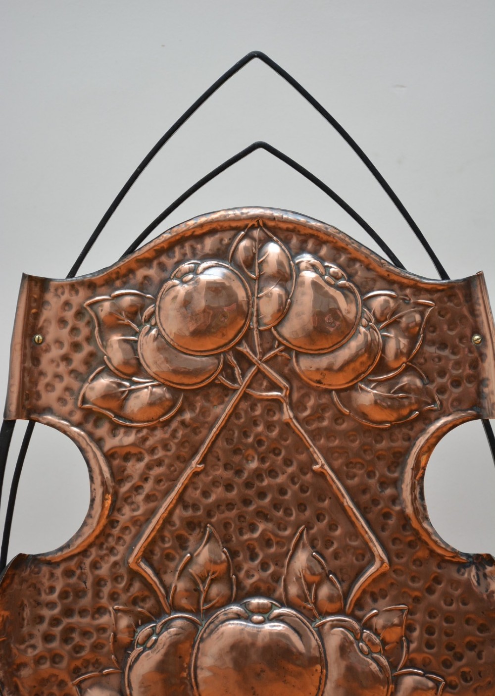 An Arts and Crafts fire screen featuring a shaped copper panel with hand-hammered botanical repousse work suspended inside a black wrought iron frame. View II