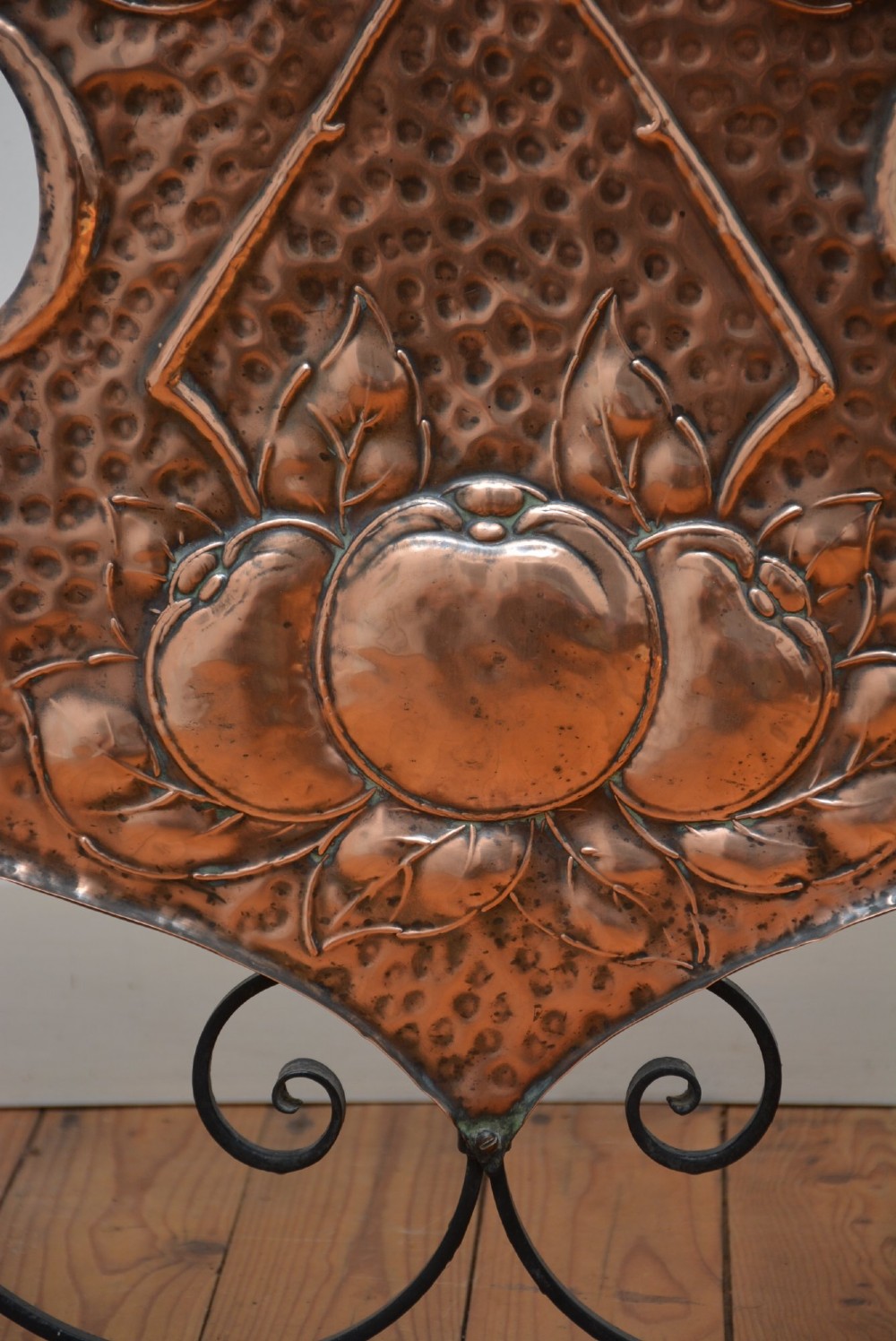 An Arts and Crafts fire screen featuring a shaped copper panel with hand-hammered botanical repousse work suspended inside a black wrought iron frame. View V