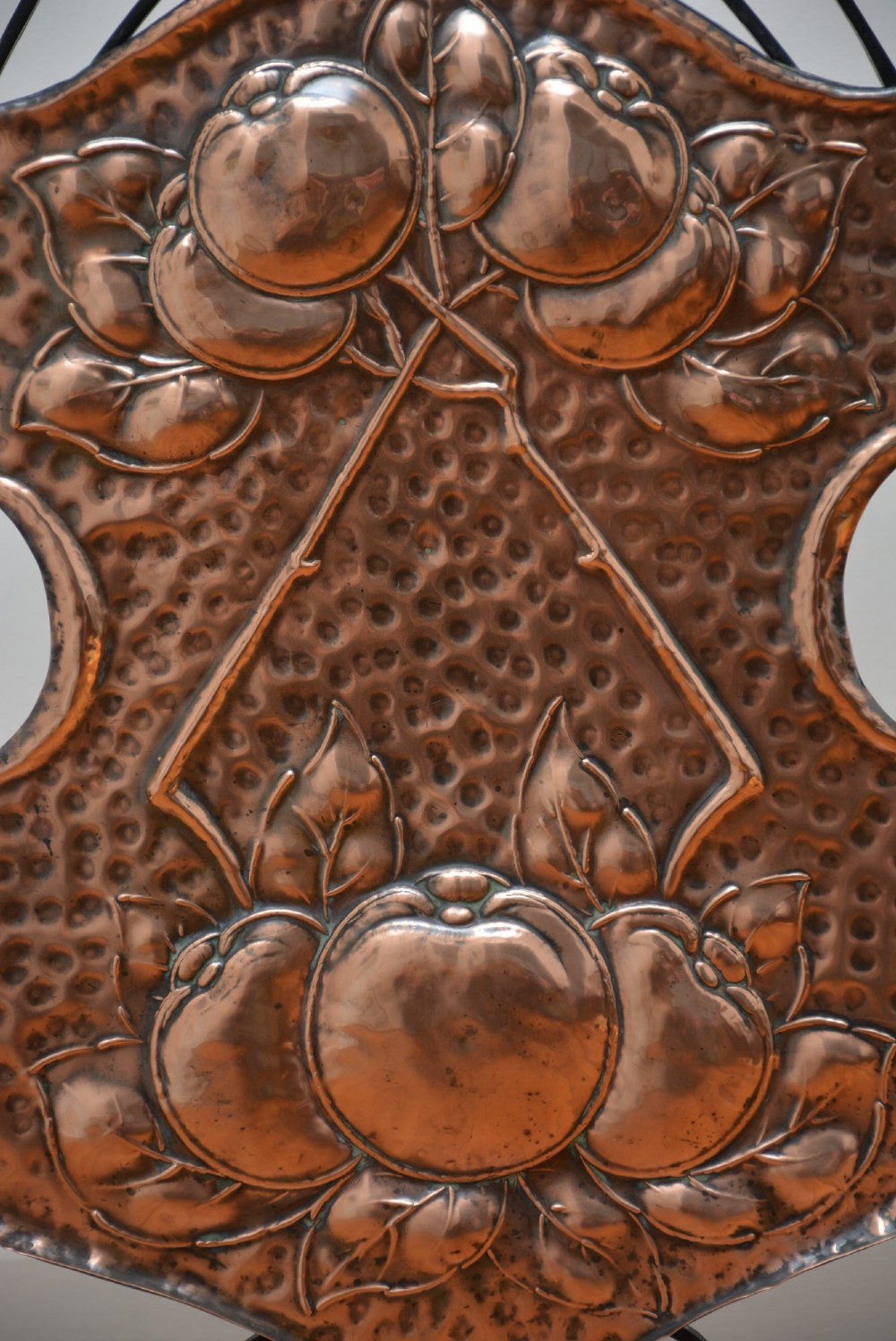 An Arts and Crafts fire screen featuring a shaped copper panel with hand-hammered botanical repousse work suspended inside a black wrought iron frame. View VI