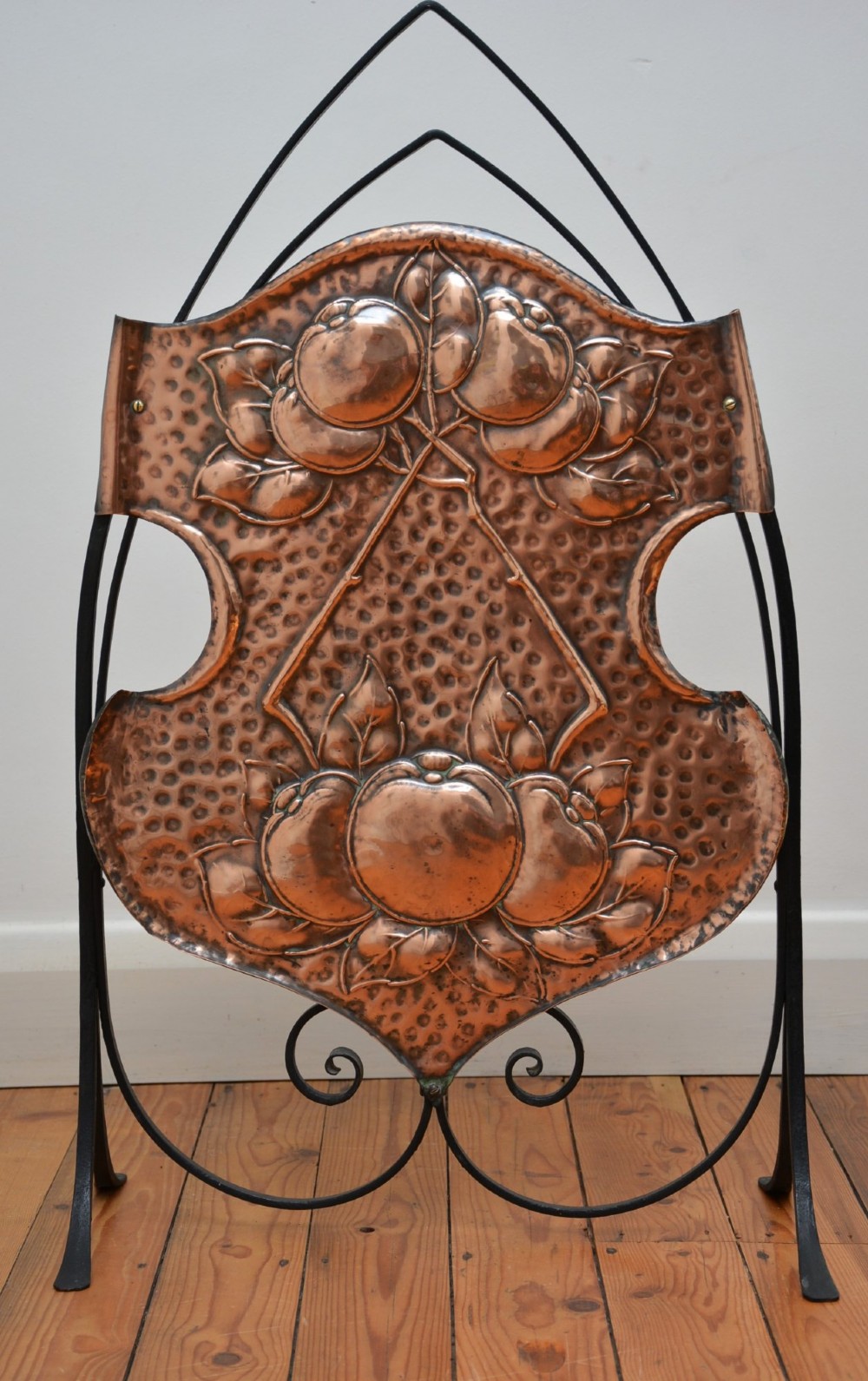 An Arts and Crafts fire screen featuring a shaped copper panel with hand-hammered botanical repousse work suspended inside a black wrought iron frame. View VII