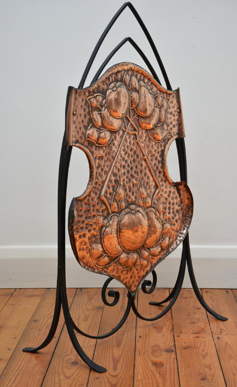 An Arts and Crafts fire screen featuring a shaped copper panel with hand-hammered botanical repousse work suspended inside a black wrought iron frame. View VIII