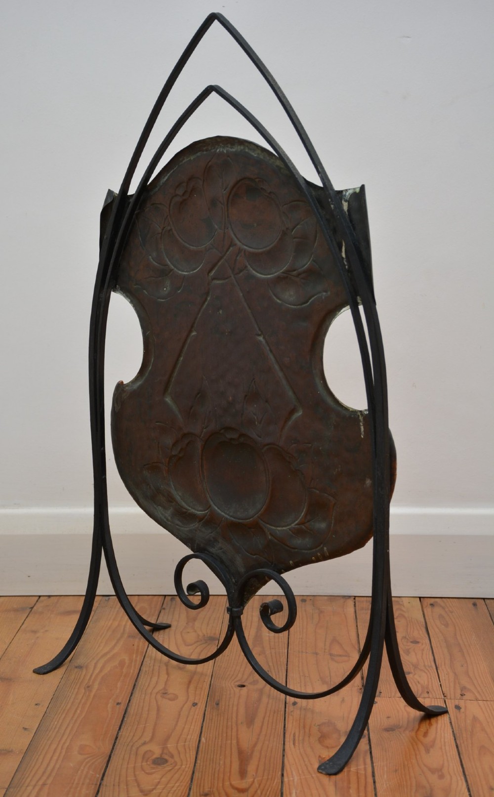 An Arts and Crafts fire screen featuring a shaped copper panel with hand-hammered botanical repousse work suspended inside a black wrought iron frame. View X