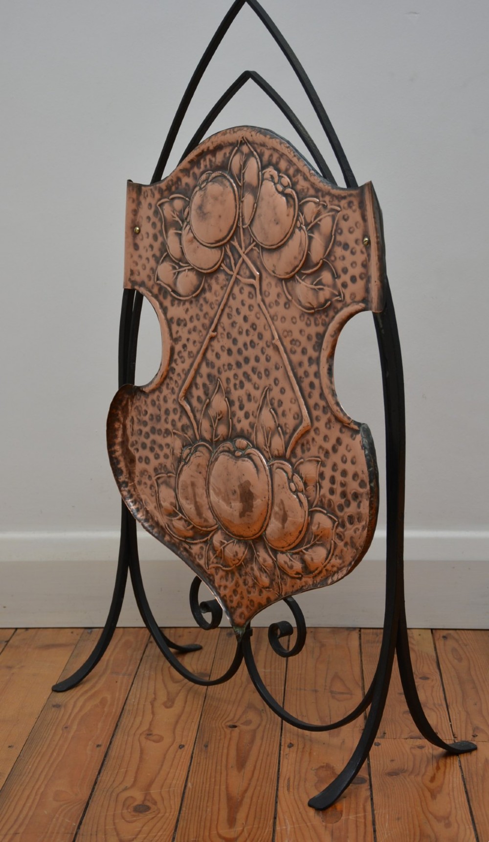 An Arts and Crafts fire screen featuring a shaped copper panel with hand-hammered botanical repousse work suspended inside a black wrought iron frame. View XII
