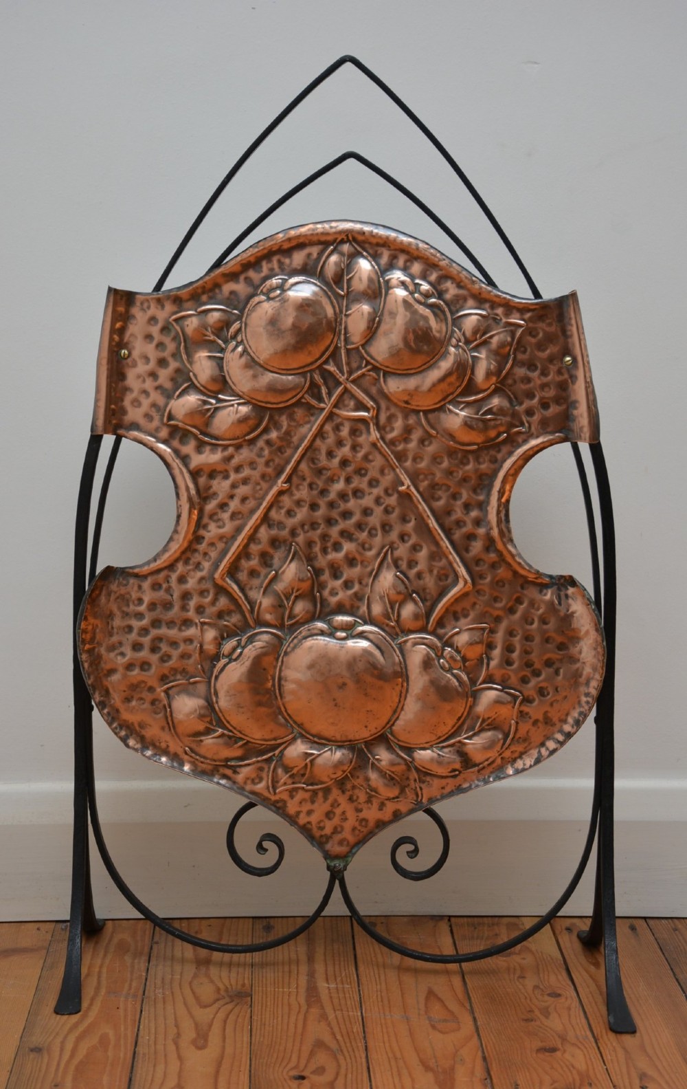 An Arts and Crafts fire screen featuring a shaped copper panel with hand-hammered botanical repousse work suspended inside a black wrought iron frame. View XIII