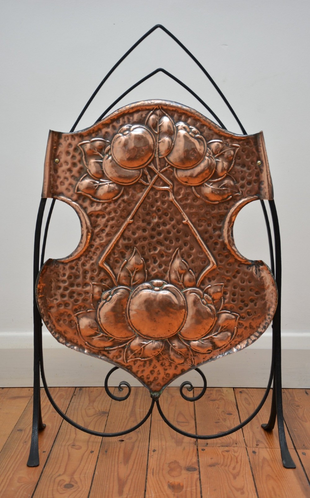An Arts and Crafts fire screen featuring a shaped copper panel with hand-hammered botanical repousse work suspended inside a black wrought iron frame.